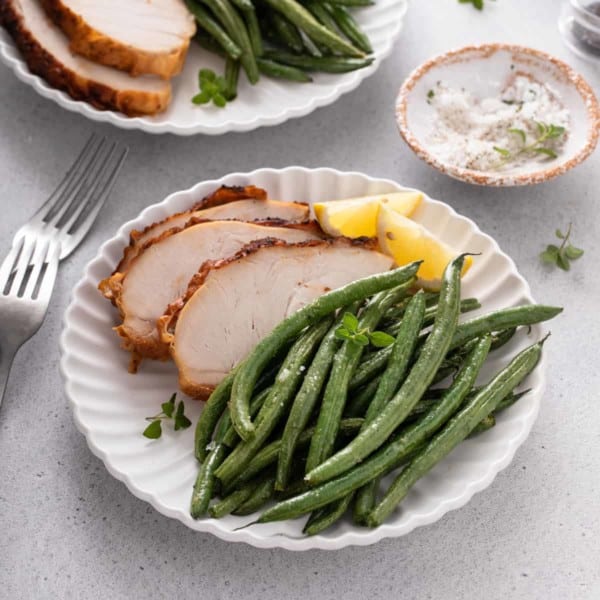 Air fryer green beans on a white plate next to roasted chicken and slices of lemon.