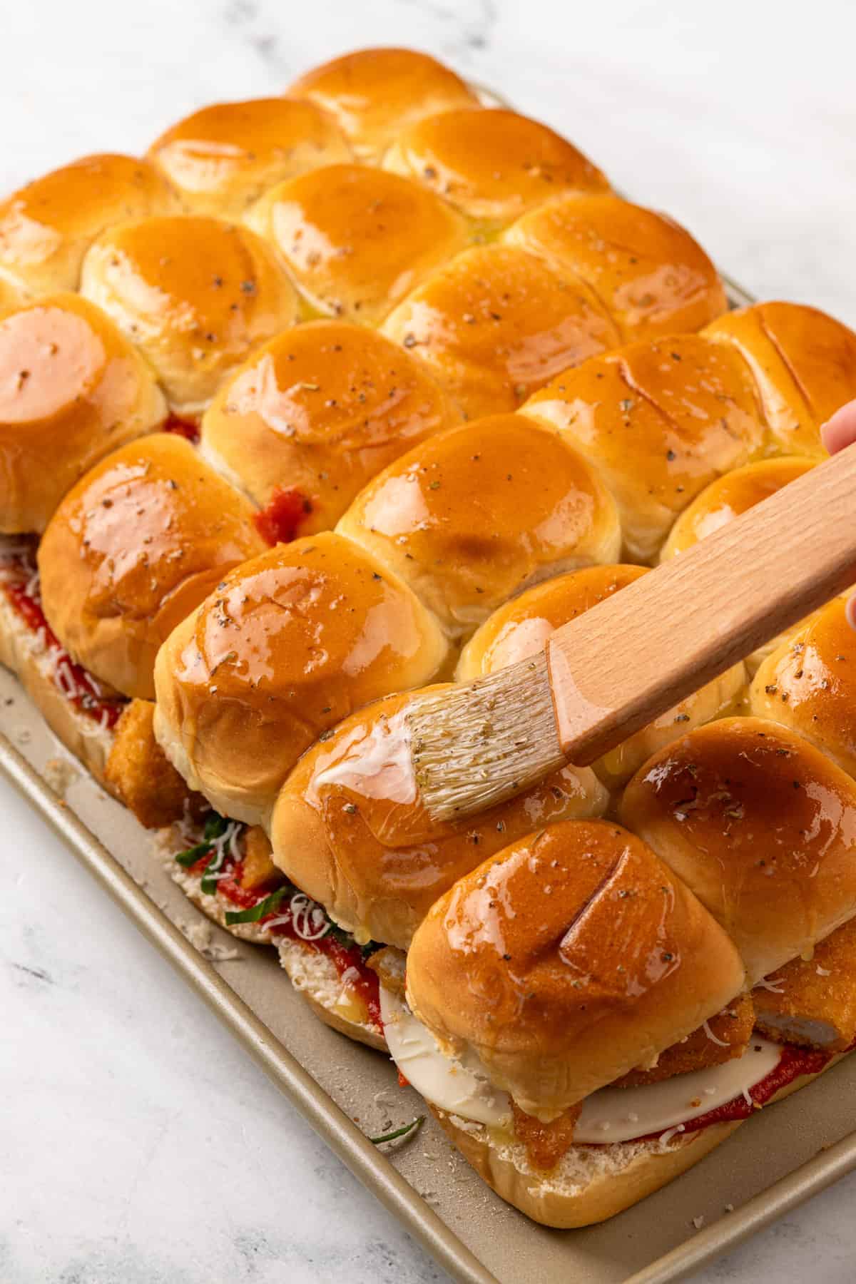 Brushing melted butter over the tops of assembled chicken parmesan sliders on a rimmed baking sheet.