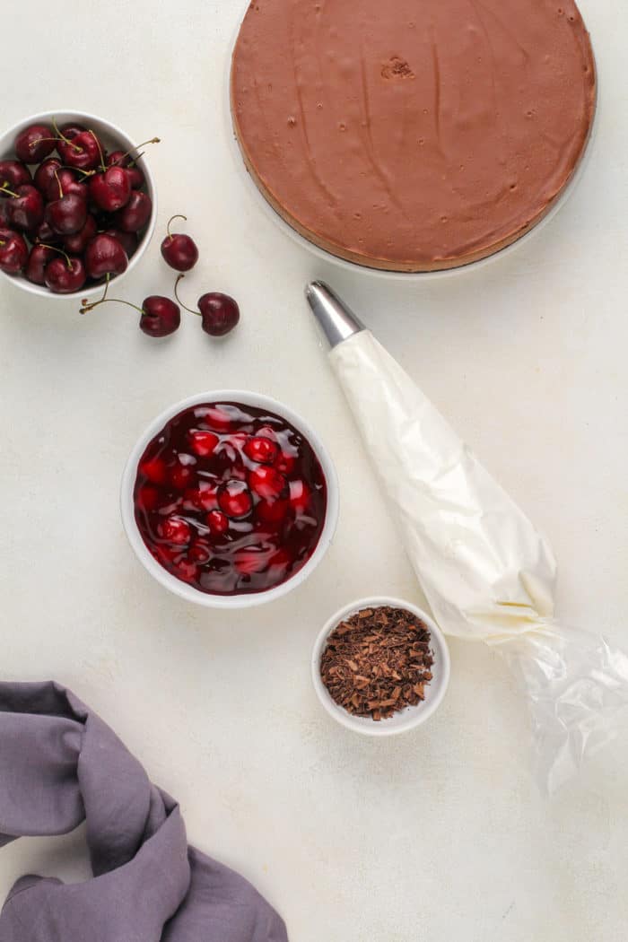 Baked chocolate cheesecake next to a bowl of cherry pie filling, whipped cream, and chocolate shavings.