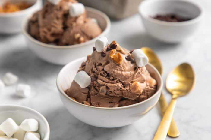 White bowl of rocky road ice cream set on a marble countertop next to gold spoons.