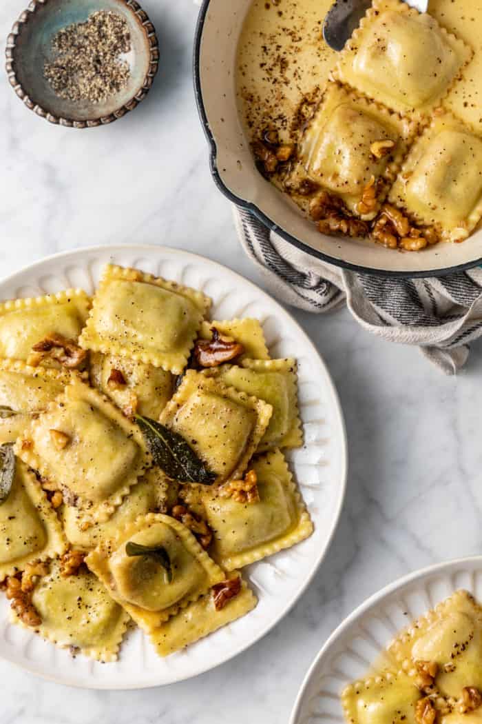 Ravioli tossed with brown butter sauce on a white plate, set on a marble countertop.