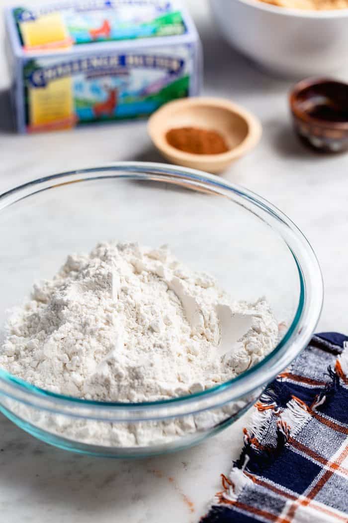 Glass bowl filled with flour in front of small bowls of spices and a package of butter