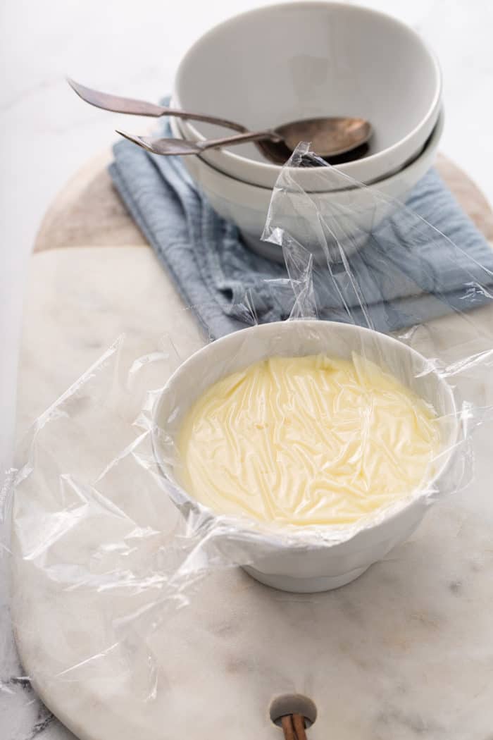 Small white bowl full of homemade vanilla pudding with plastic wrap pressed onto the top of the pudding