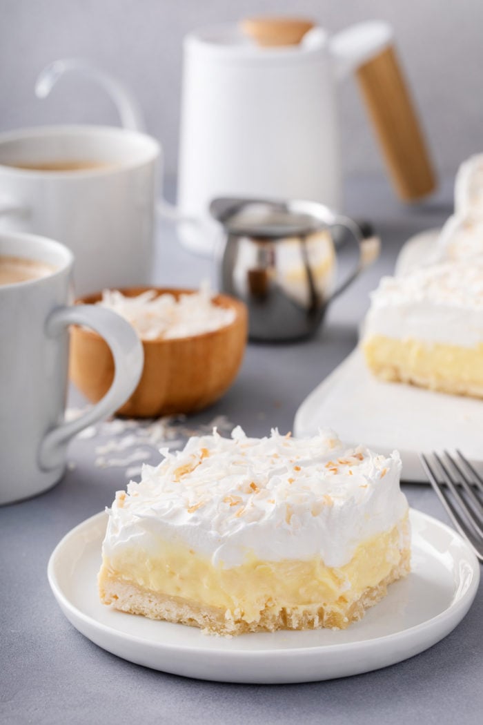 Plated coconut cream bar with cups of coffee in the background. A bite has been taken from the corner of the bar.