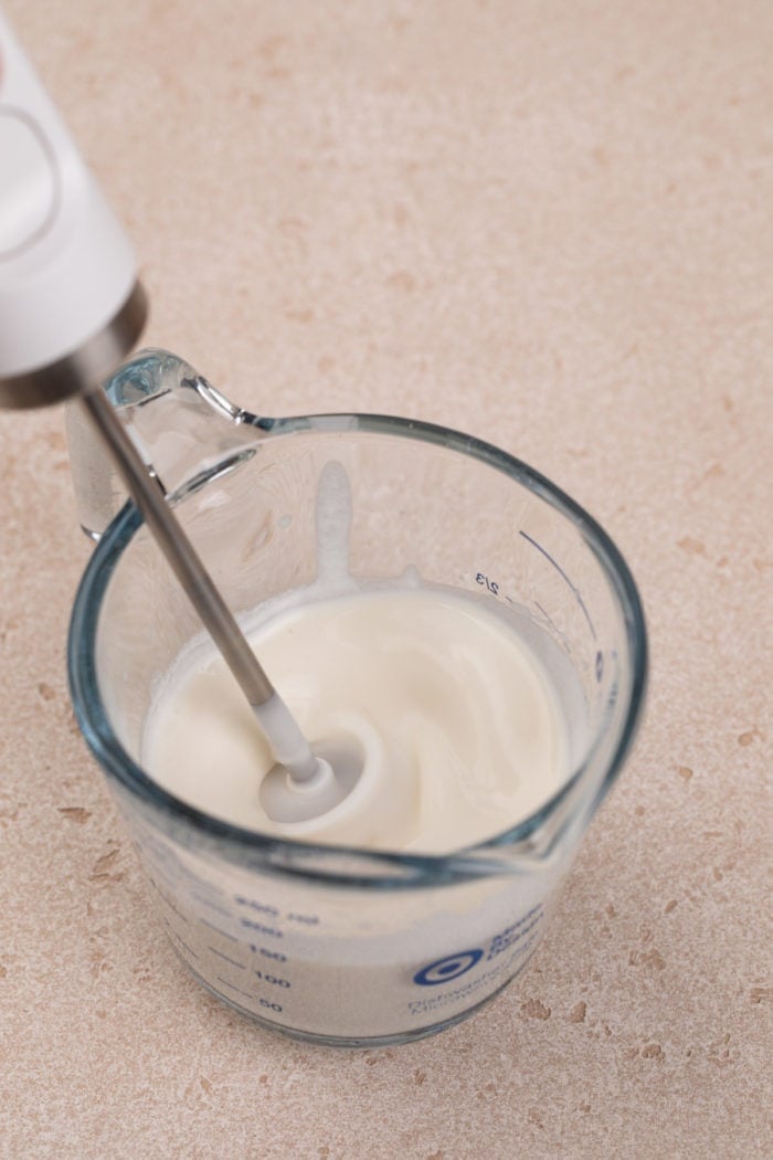 Vanilla sweet cream cold foam being frothed with a hand frother in a glass measuring cup.