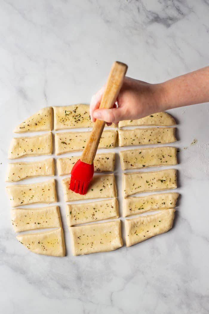 Hand using a pastry brush to brush herb butter onto rolled and cut dough for garlic and herb parker house rolls