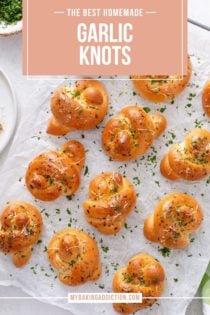 Overhead view of baked garlic knots on a piece of parchment paper, garnished with parsley and parmesan cheese. Text overlay includes recipe name.