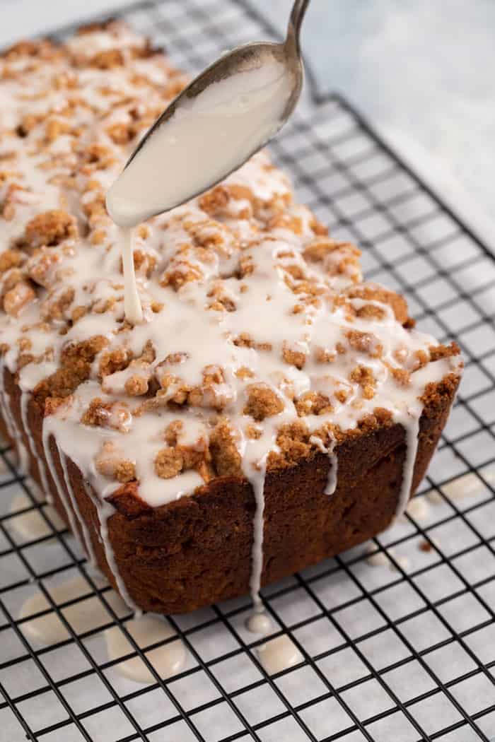 Spoon drizzling glaze over a loaf of apple fritter bread on a wire rack