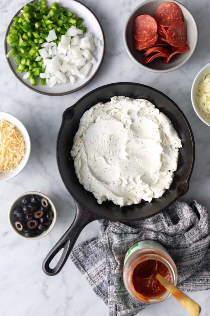 Herbed cream cheese spread into the bottom of a cast iron skillet. The rest of the ingredients for pizza dip are in bowls around the skillet.