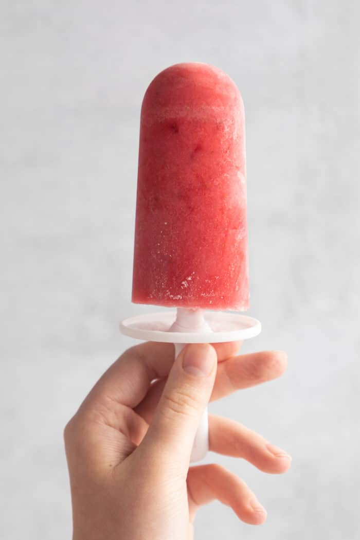 Hand holding a strawberry popsicle in front of a white wall.