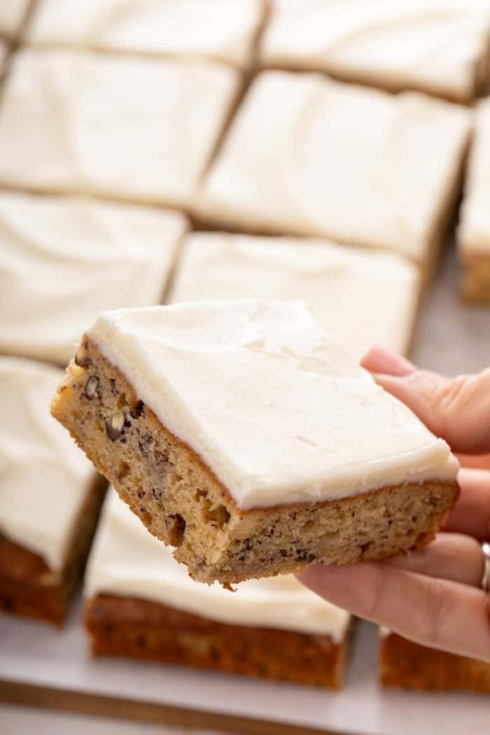 Hand holding a frosted banana bar up to the camera; the pan of sliced bars is visible in the background.