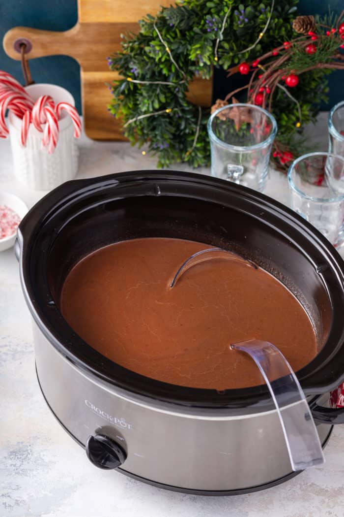 Large slow cooker filled with hot chocolate on a countertop, with glasses and toppings in the background.