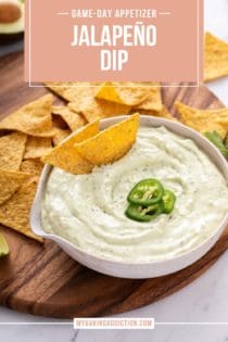 White bowl filled with jalapeño dip, with two chips in the dip. The bowl is on a wooden platter surrounded by tortilla chips. Text overlay includes recipe name.