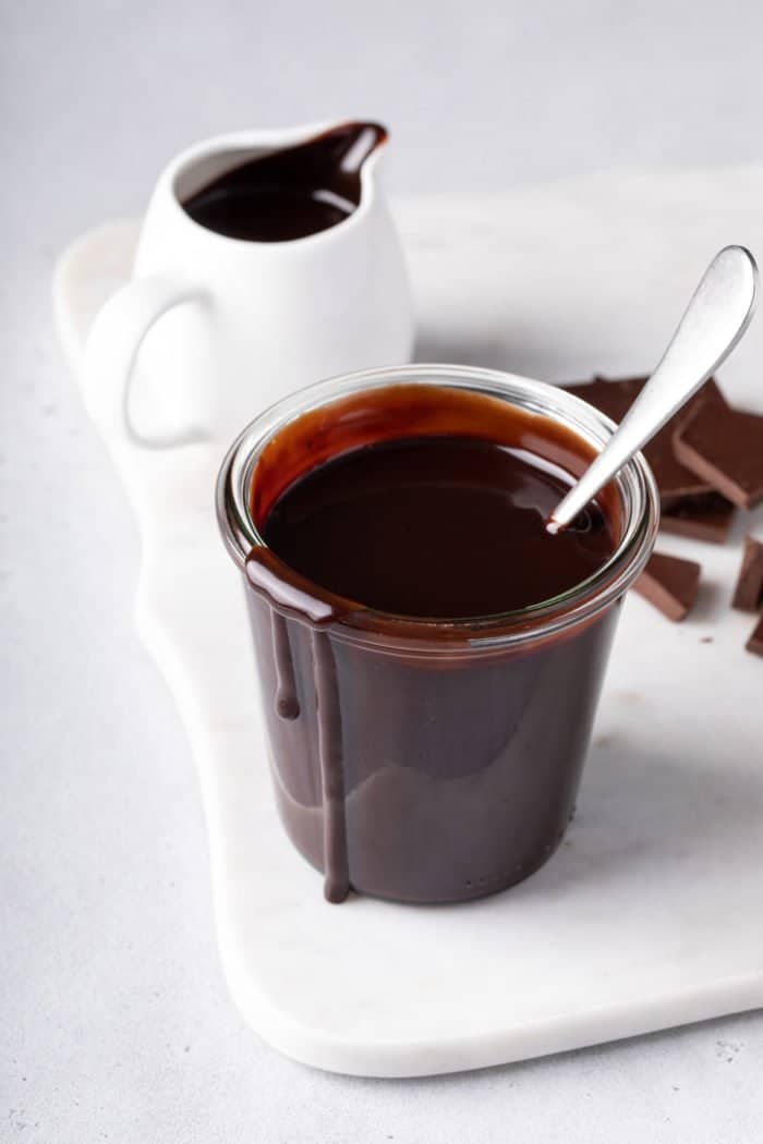 Spoon resting in a glass jar of hot fudge sauce set on a white board.