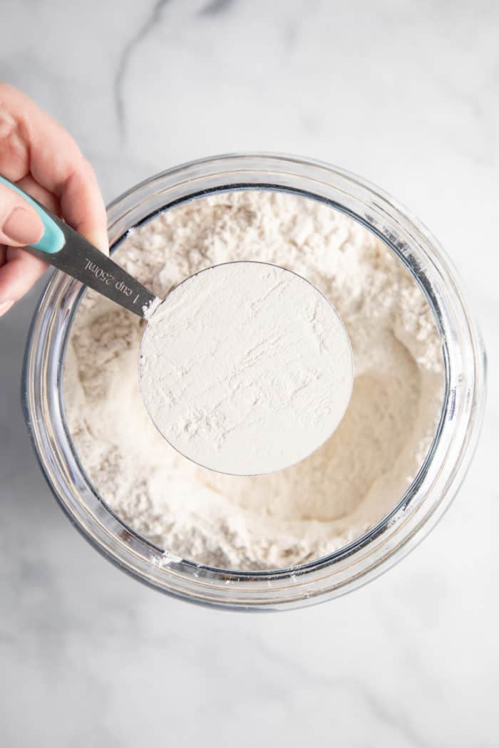 Leveled measuring cup of flour held over a glass bowl of flour.