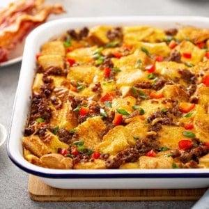 Baking dish of make-ahead breakfast casserole on a wooden trivet with a plate of cooked bacon in the background