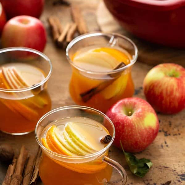 Three mugs of hot wassail garnished with sliced apples and cinnamon sticks with a dutch oven full of wassail in the background