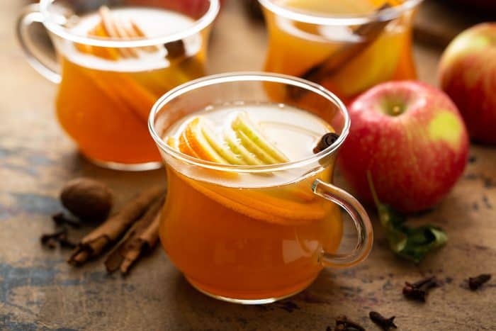 Mug of wassail with apples, cinnamon sticks, and two more mugs of wassail in the background