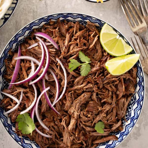 Overhead view of chipotle barbacoa in a blue and white bowl, toped with red onions, cilantro and lime wedges