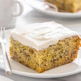 Slice of frosted moist banana cake on a white cake next to a silver fork