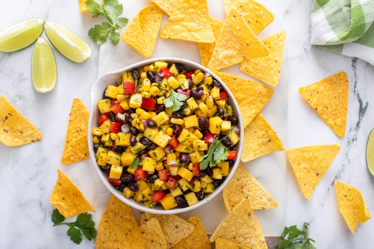 Overhead view of a bowl of mango and black bean salsa on a countertop, with chips and lime wedges scattered around it.