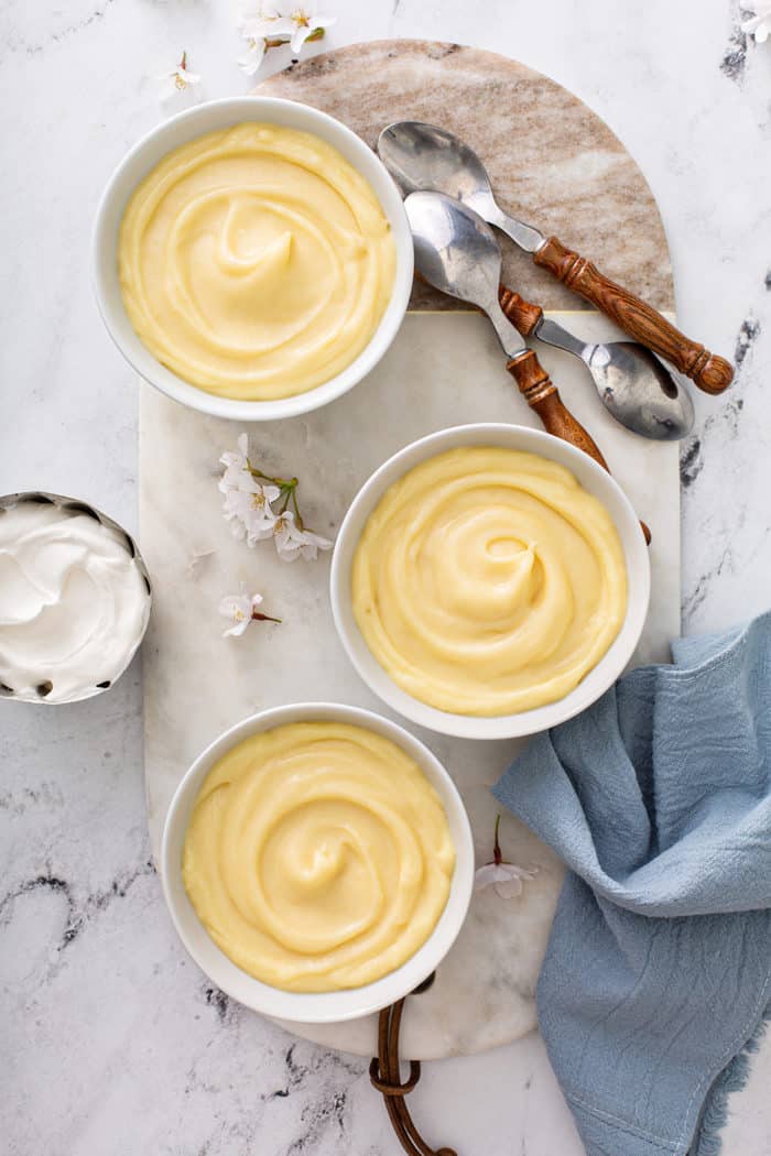 Overhead view of three white bowls of homemade vanilla pudding on a marble board, surrounded by spoons and homemade whipped cream