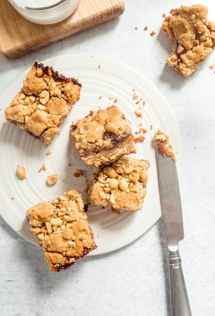 Four peanut butter and jelly bars scattered on a white plate next to a knife with peanut butter on the tip