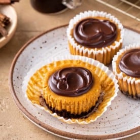 Unwrapped mini pumpkin cheesecake set on a plate next to two other mini pumpkin cheesecakes.