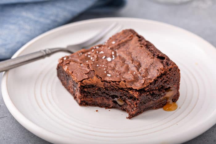 Salted caramel brownie next to a fork on a white plate, with a bite taken out of it. Caramel is pooling out of the edge of the brownie