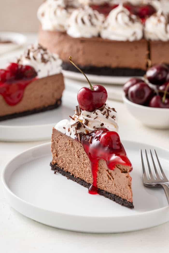 Slice of garnished black forest cheesecake next to a fork on a white plate.