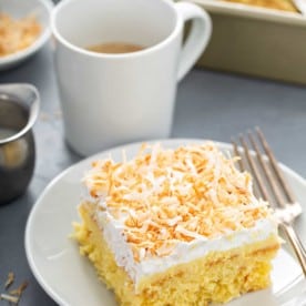 Slice of coconut poke cake next to a fork on a white plate, with a cup of coffee in the background