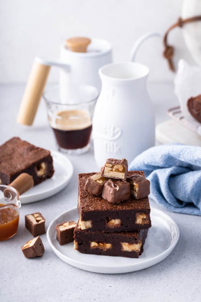 Two snickers brownies stacked on a white plate, topped with pieces of snickers bars.