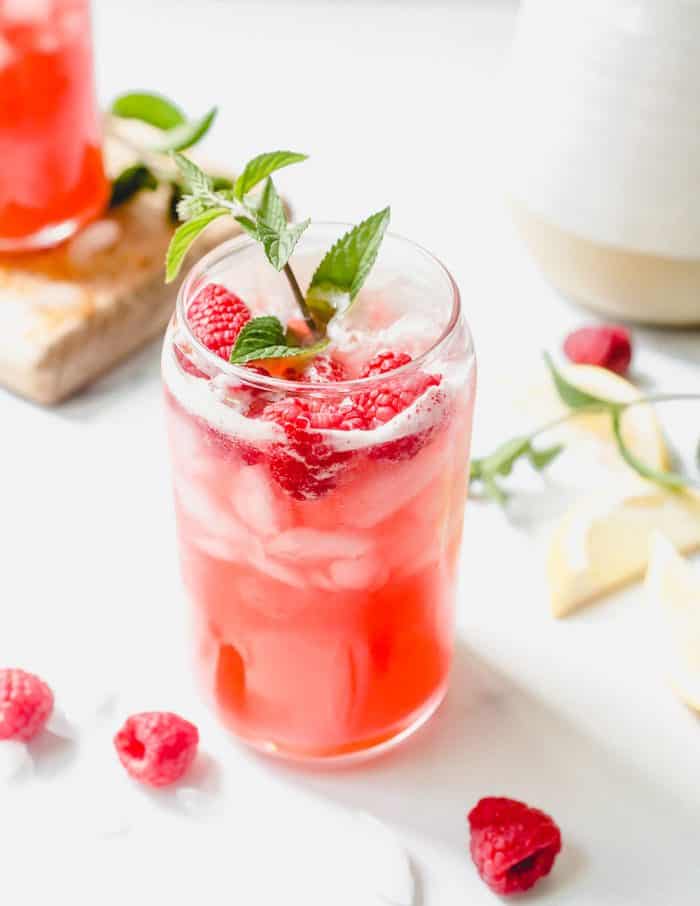 Glass of raspberry lemonade garnished with a sprig of mint on a white counter, with raspberries scattered on the countertop