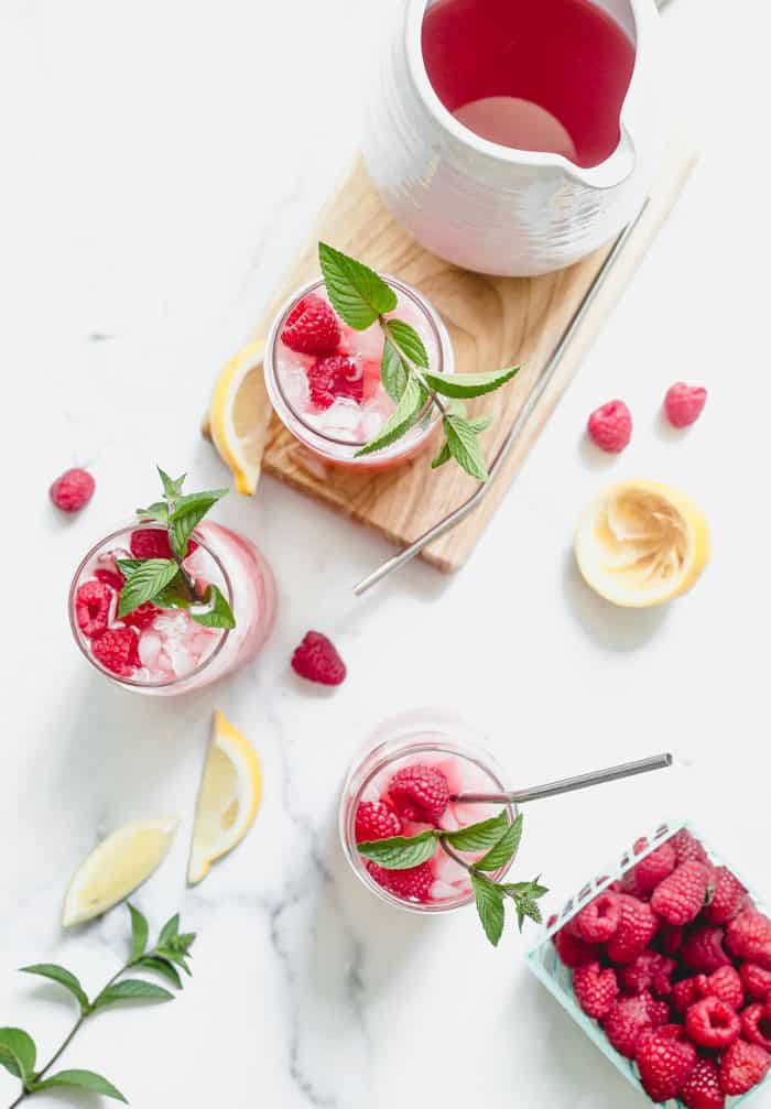 Overhead view of three glasses of raspberry lemonade on a white counter, surrounded by lemon slices and fresh raspberries