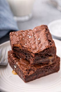 Two salted caramel brownies stacked on a white plate. The top brownie has a bite taken out of it and caramel is pooling out of the edges of the brownies