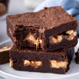 Two snickers brownies stacked on a plate. A bite has been taken out of the corner of the top brownie.