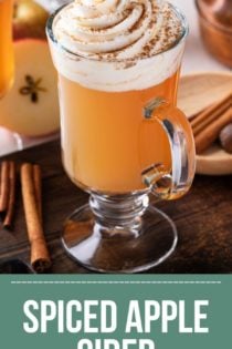 Mug of spiced apple cider on a wooden board. Two more mugs of cider are visible in the background. Text overlay includes recipe name.