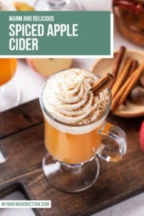 45-degree overhead view of a glass mug of spiced apple cider, topped with whipped cream and garnished with a cinnamon stick. Text overlay includes recipe name.