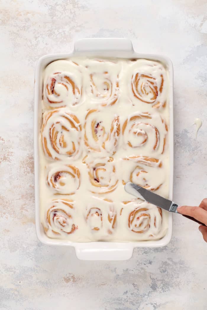 Hand spreading cream cheese frosting onto strawberry rolls with an offset spatula.