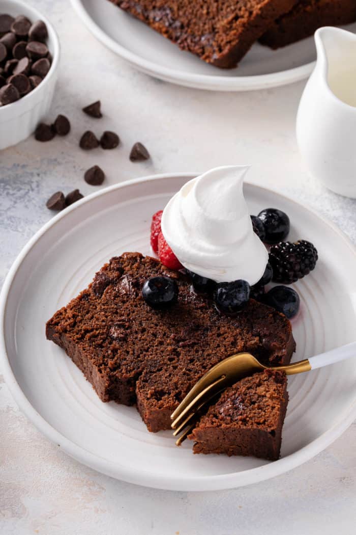 Fork taking a bite from a piece of chocolate bread on a white plate, topped with whipped cream and berries.