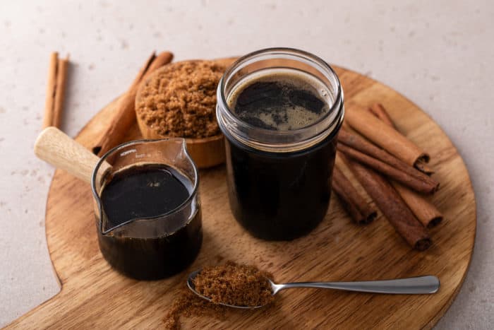 Two jars filled with brown sugar syrup set on a wooden board and surrounded by brown sugar and cinnamon sticks.