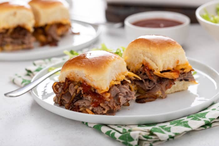 White plate with two cheesy roast beef sliders on it, with a second plate in the background.