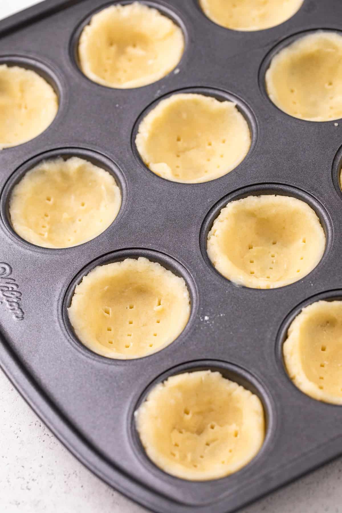 Tart dough pressed into mini muffin wells and poked with a fork, ready to go in the oven.