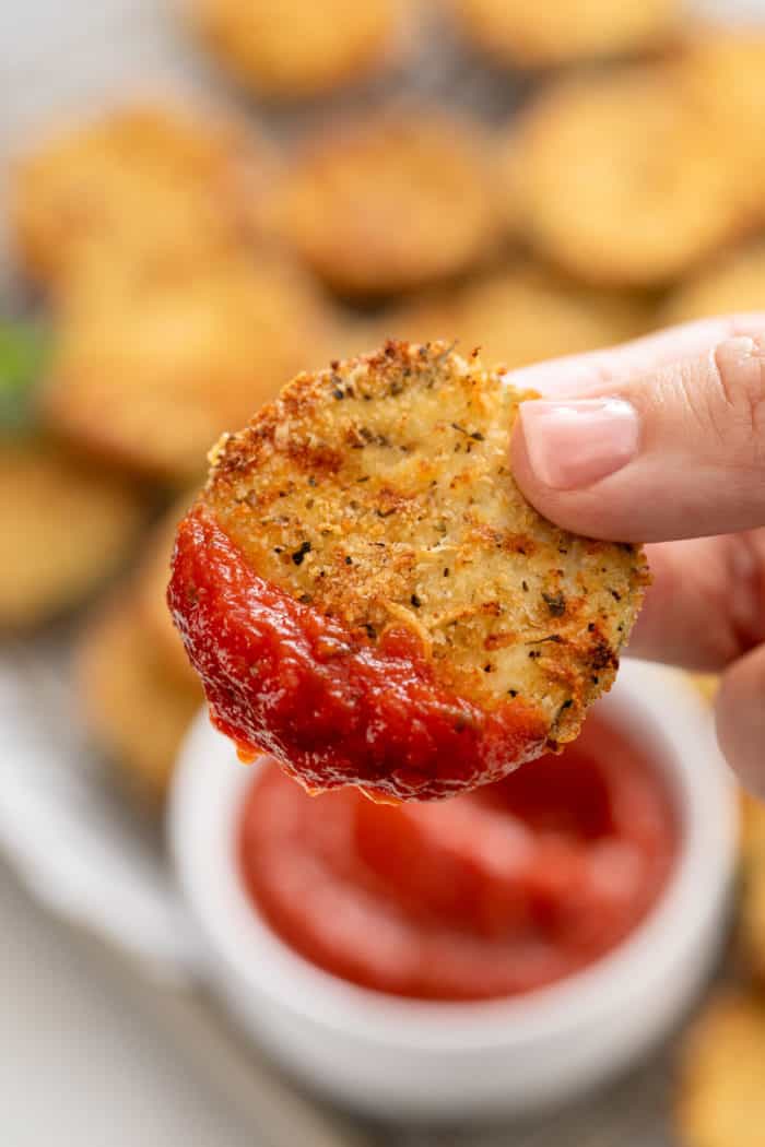 Hand holding up an air fryer zucchini chip dipped in marinara sauce.